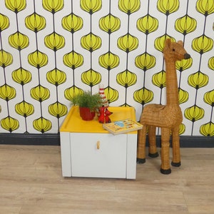 Può includere: Un mobile bianco con un piano giallo e un solo cassetto. Il mobile è appoggiato su un pavimento di legno davanti a una parete con un motivo geometrico giallo e bianco. Una statuetta di giraffa in vimini è in piedi accanto al mobile.