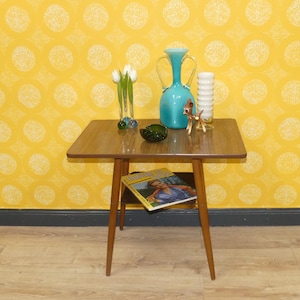 Könnte beinhalten: Ein Mid-Century-Modern-Beistelltisch mit quadratischer Tischplatte und konischen Beinen. Auf dem Tisch stehen eine türkisfarbene Vase, ein weißer Kerzenhalter, eine kleine Hirschfigur, ein grüner Aschenbecher und eine Zeitschrift. Der Hintergrund ist eine gelbe Tapete mit Muster.