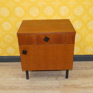 Könnte beinhalten: Ein Nachttisch aus Holz im Vintage-Stil mit zwei Schubladen und schwarzen Rauten-Details. Der Schrank hat eine warme braune Oberfläche und steht auf schwarzen Metallbeinen. Das Design ist schlicht und funktional, passend für ein Schlaf- oder Wohnzimmer. Höhe ca. 60 cm.