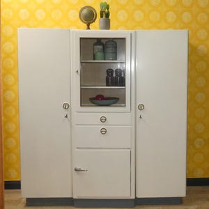 Könnte beinhalten: Ein weißer Vintage-Schrank mit einem zentralen Abschnitt mit Glasfront, Schubladen und Türen. Der Schrank hat einen dunkelgrauen Sockel und ist mit dekorativen Gegenständen wie einem Globus und einem kleinen Kaktus versehen. Der Schrank steht vor einer gelben Tapete.