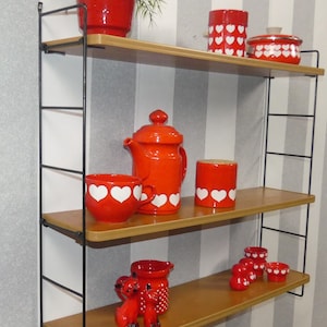 May include: A black metal shelf unit with three wooden shelves. The shelves are decorated with red and white ceramic items, including a teapot, a bowl, a jar, and a pitcher. There is also a small potted plant with green leaves.