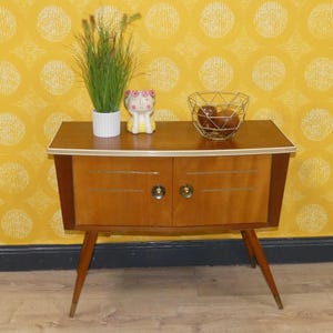 Puede incluir: Un mueble de madera de estilo vintage con dos puertas y tiradores de latón. El mueble tiene un acabado marrón claro con un ribete dorado. Una pequeña maceta blanca con hierba verde, una figura decorativa y una cesta de alambre dorada con fruta encima. Mide 75 cm de ancho.