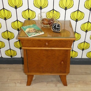 Könnte beinhalten: Ein Nachttisch aus Holz mit einer Schublade und einer Schranktür. Der Tisch hat eine Glasplatte und steht auf vier Beinen. Ein Buch mit dem Titel "Lotje" und ein kleiner Wecker stehen auf dem Tisch.