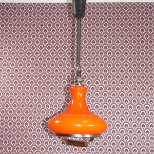 May include: Orange glass pendant light with a black cord and a metal ceiling mount. The light is hanging from a ceiling with a patterned wallpaper.