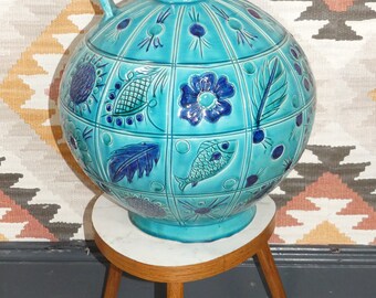 Può includere: Un grande vaso rotondo in ceramica turchese con manico e intricati disegni floreali e di pesci blu e verdi. Il vaso è posto su un piedistallo in legno a tre gambe.