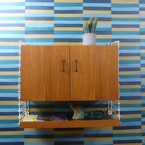 Op de afbeelding: Een wandrek bestaande uit een houten kast met twee deuren en zwarte handgrepen, ondersteund door witte metalen draadpanelen. Onder de kast bevindt zich een open houten plank met een klein boek en een groen glazen schaaltje.