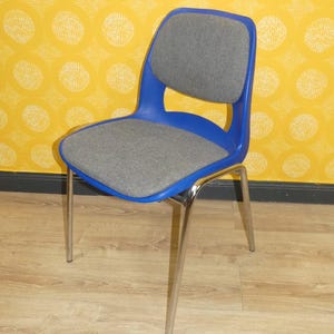 May include: A blue and gray chair with chrome legs. The chair has a blue plastic frame with a gray upholstered seat and back. The legs are made of shiny chrome. The chair is set against a yellow patterned wall.