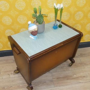 Könnte beinhalten: Ein Vintage-Beistelltisch aus Holz mit hellgrüner Oberseite und braunem Rahmen. Der Tisch hat ein geschwungenes Design, kleine Räder und einen Griff an der Seite. Dekoriert mit einem Kaktus, einer Figur und Vasen.