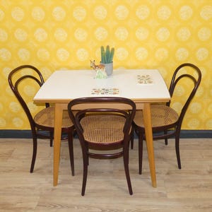 Könnte beinhalten: Ein kleines Esszimmer-Set mit einer weißen Tischplatte und vier dunkelbraunen Stühlen mit gewebten Sitzen. Die Stühle sind um den Tisch angeordnet. Im Hintergrund befindet sich eine gelbe Tapete mit Muster. Dekorative Gegenstände befinden sich auf dem Tisch.