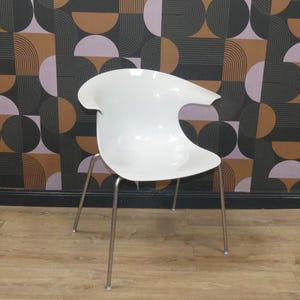 May include: A white moulded plastic chair with a curved, shell-like seat and integrated armrests. The chair is supported by four slender, straight metal legs with a silver finish, providing a stable base for the smooth, glossy white seat structure.