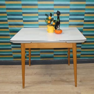 Könnte beinhalten: Ein rechteckiger Tisch mit weißer Tischplatte und hellbraunen Holzbeinen. Auf dem Tisch stehen ein gelber Topf mit einer kleinen Pflanze mit orangefarbenen Früchten, eine schwarze Katzenfigur und eine kleine orangefarbene Schale mit silbernem Deckel.