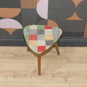 May include: A triangular side table featuring a tabletop with a multi-coloured grid pattern of red, yellow, green, and grey square tiles. The table has a thin gold-toned metal rim around the edge and three tapered wooden legs extending downwards.