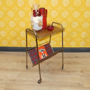 May include: A vintage gold-toned serving cart with two tiers. The top tier holds a red pitcher, a small red bowl, and a wooden Viking figurine. The bottom tier has a magazine rack with a plaid pattern and a magazine with a cowboy on the cover.