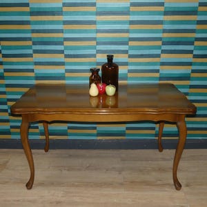 Könnte beinhalten: Ein rechteckiger Couchtisch aus Holz mit geschwungenen Beinen und glänzender Oberfläche. Zwei braune Glasflaschen, eine Birne und zwei Äpfel stehen auf dem Tisch. Der Hintergrund zeigt eine Wand mit horizontalen Streifen in Türkis, Blau und Gold.