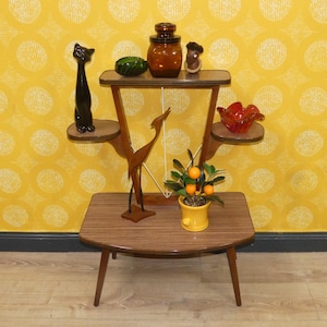 May include: A mid-century modern plant stand with multiple shelves, holding various decorative items. A black cat figurine, a green glass dish, a brown jar, and a small wooden figure are displayed. A wooden bird sculpture and a yellow pot with orange-like fruit complete the arrangement.
