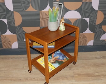 Puede incluir: Un carrito de madera con ruedas, dos estantes y cuatro rodines. El estante superior sostiene una maceta gris con una suculenta verde, un jarrón blanco alto con patrones negros y una pequeña figura de ciervo. El estante medio tiene un libro y el inferior una revista.
