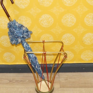 Puede incluir: Un paragüero vintage con estructura metálica dorada y detalles rojos. Un paraguas azul con estampado floral y mango curvo de madera se encuentra dentro. El fondo es una pared amarilla con un patrón circular blanco.