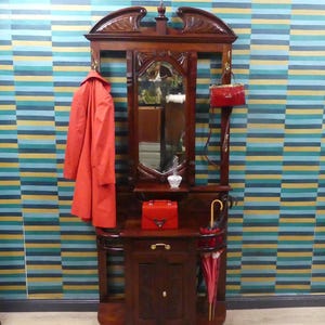 Könnte beinhalten: Eine Garderobe aus dunklem Holz mit einem zentralen Spiegel, einer kleinen Ablage, einem Unterschrank mit Schublade und seitlichen Schirmständern. Ein leuchtend orangefarbener Mantel hängt links, während eine rote Handtasche und ein roter Schirm präsentiert werden.