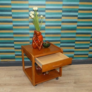 Könnte beinhalten: Ein Beistelltisch aus Holz im Mid-Century-Modern-Stil mit einer geöffneten Schublade. Der Tisch hat ein unteres Regal und steht auf kleinen Rollen. Eine Vase mit einer Skulptur und eine grüne Schale stehen oben.