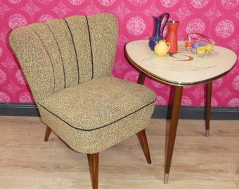 Op de afbeelding: Een moderne stoel uit het midden van de eeuw met een bruine stoffen bekleding en een zwarte bies. De stoel heeft een gebogen rugleuning en houten poten. Een kleine, driehoekige tafel met een wit blad en houten poten staat naast de stoel. De tafel heeft een decoratief ontwerp op het blad en is versierd met drie kleine vazen.