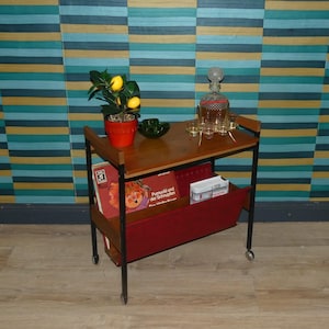 Può includere: Un carrello bar vintage con piano in legno e portariviste rosso. Contiene una pianta di limone in vaso, una caraffa, bicchieri e libri. Il carrello ha una struttura in metallo nero e ruote.