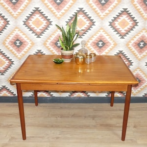 Könnte beinhalten: Ein rechteckiger Esstisch aus Holz mit einer hellen Holzoptik und einer geometrisch gemusterten Tapete im Hintergrund. Der Tisch hat eine Auszugsfläche und vier Beine. Auf dem Tisch stehen eine Topfpflanze, ein goldfarbenes Dekoobjekt und zwei Kerzenhalter aus Glas.