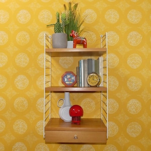 May include: A three-tiered wooden shelf unit with white metal supports, displaying decorative items. Includes a cactus, a red horse figurine, books, a clock, a vase, and a red mushroom-shaped lamp. The background is yellow wallpaper with a white circular pattern.