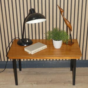 Könnte beinhalten: Ein Holz-Beistelltisch mit schwarzen Metallbeinen. Auf dem Tisch liegen ein Buch, eine Topfpflanze und eine Holzvogelskulptur.
