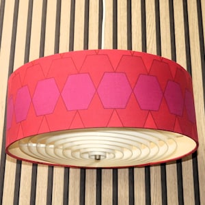 Puede incluir: Una lámpara colgante con un patrón geométrico rojo y rosa. La pantalla tiene un patrón repetitivo de hexágonos y diamantes rosas sobre un fondo rojo. La luz tiene un interior blanco, en capas y en forma de espiral.