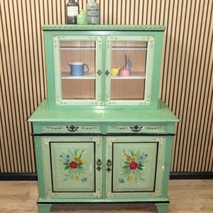 Könnte beinhalten: Ein grüner, bemalter Holzschrank mit zwei Glastüren oben und zwei massiven Türen unten. Der Schrank hat zwei Schubladen und florale Bemalungen auf den Türen. Der Schrank steht auf einem gestreiften Hintergrund.
