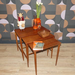 Könnte beinhalten: Ein Set aus drei ineinander stapelbaren Holztischen mit konisch zulaufenden Beinen. Auf den Tischen stehen eine Wikingerfigur, zwei orangefarbene Keramikvasen mit grünen Stielen, eine Holzschale und ein Buch mit blauem Einband namens The American Way of Life.