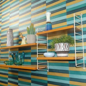 May include: A modular wall shelving unit with white metal wire side supports and three light brown wooden shelves. The shelves hold various decorative items including a gray vase, a small deer figurine, a green faux cactus, a blue glass bowl, and a patterned planter.
