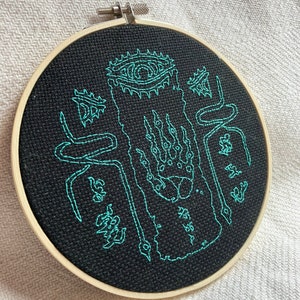 Może przedstawiać: Czarne tamborek do haftu z zielonym haftem przedstawiającym stworzenie z okiem, pazurami i wężami. Wz&oacute;r znajduje się na czarnym tle tkaniny.