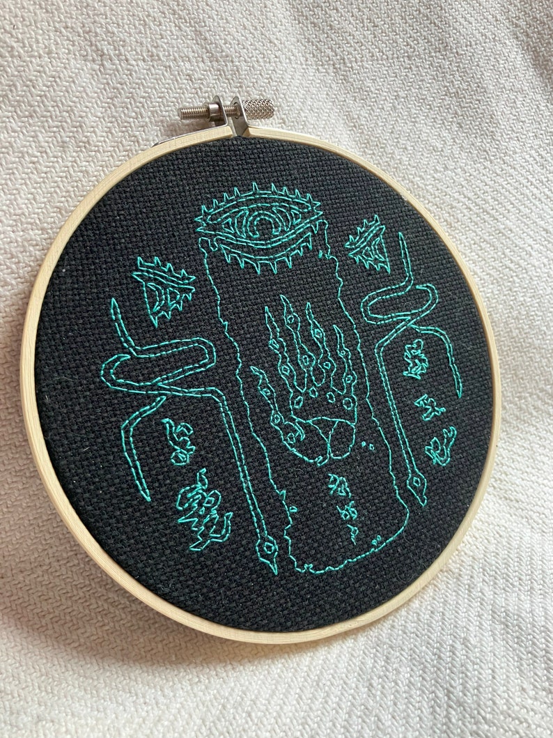 Może przedstawiać: Czarne tamborek do haftu z zielonym haftem przedstawiającym stworzenie z okiem, pazurami i wężami. Wz&oacute;r znajduje się na czarnym tle tkaniny.