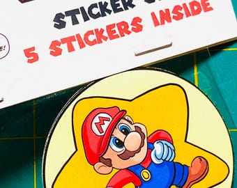 May include: A sticker set with the text "LEVEL UP DANCE!" and "STICKER SET 5 STICKERS INSIDE." The set includes a Mario sticker inside a yellow star and a frog character. The stickers are on a green cutting mat.