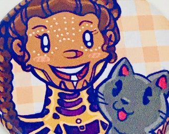 May include: A round button featuring a cartoon illustration of a person with brown skin and brown hair, wearing a purple and gold outfit, and a gray cat with pink ears. The background is a yellow and white checkered pattern.