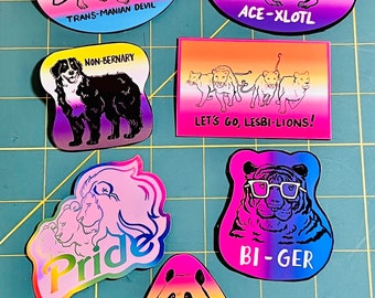 May include: A set of ten colorful stickers featuring various animals and pride flag colors. The stickers include a tiger with glasses, a panda, a lion, a dog, a quoll, and an axolotl. The stickers have text that reads "Pride", "Pan-da", "Bi-ger", "Non-binary", "Let's go, Lesbi-lions!", "Ace-Xlotl", and "Trans-manian Devil".