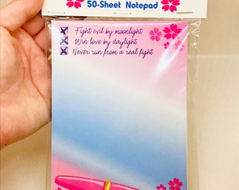 May include: A pink and gold magic wand with a red gem on top. The wand is on a pink and blue notepad with the text "Mahou Shoujo Notepad" and "50-Sheet Notepad". The notepad also has the text "Fight evil by moonlight, Win love by daylight, Never run from a real fight".