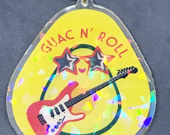 May include: A yellow avocado-shaped keychain with a green outline and a red electric guitar with a black pickguard. The avocado is wearing sunglasses with star-shaped lenses and the text "GUAC N' ROLL" is written around the top of the avocado.