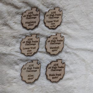 May include: Six wooden plaques with the text "ADK 46 High Peaks Challenge" and the name of a mountain peak with its elevation in feet. The plaques are arranged in two rows of three.