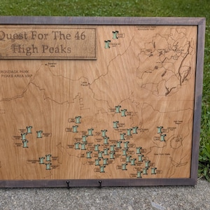 May include: Wooden map of the Adirondack High Peaks region, framed in a dark gray. The map is etched with the text "Quest For The 46 High Peaks" and includes small green mountain icons marking peaks. The map is a decorative item.