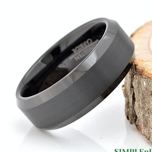 May include: A black tungsten carbide ring with a beveled edge and a brushed finish. The ring is on a wooden surface.