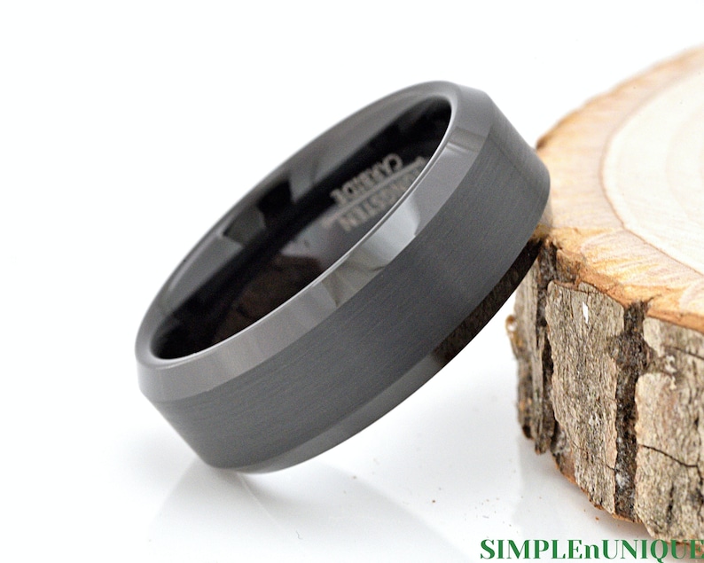 May include: A black tungsten carbide ring with a beveled edge and a brushed finish. The ring is on a wooden surface.