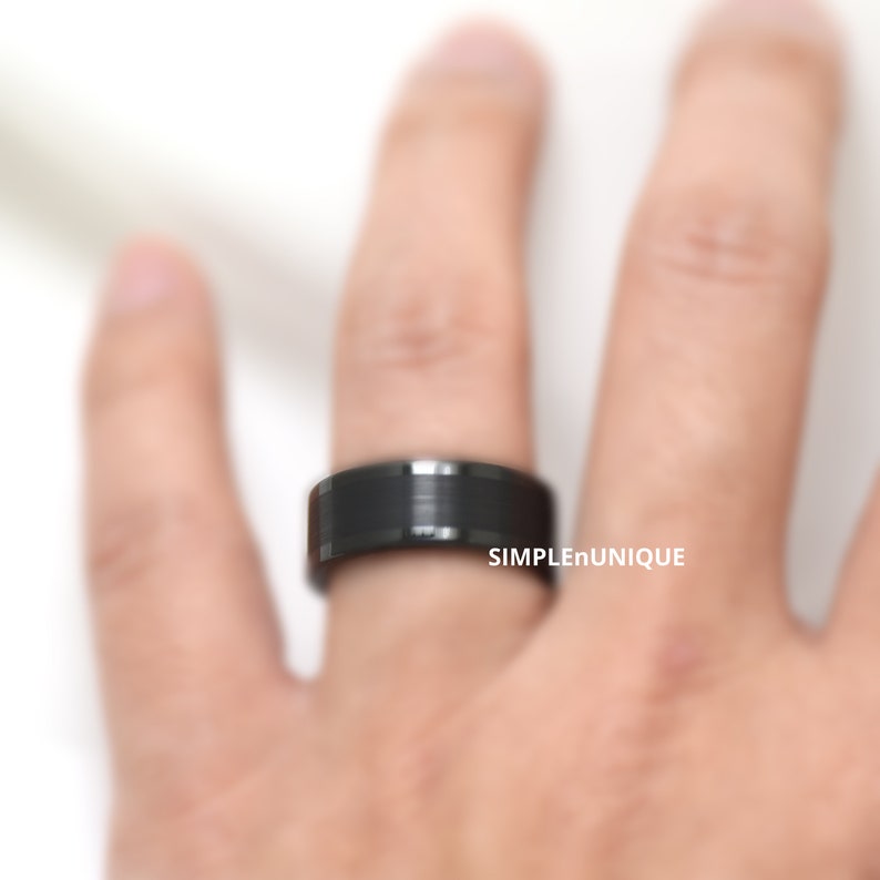 May include: A black tungsten carbide ring with a brushed finish. The ring is worn on a finger.