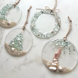May include: Four clear, circular ornaments with beach-themed designs. One features a Christmas tree, another a palm tree, and two have a half-circle of sea glass. Each has sand, shells, and a twine loop for hanging. The ornaments are approximately 3 inches in diameter.