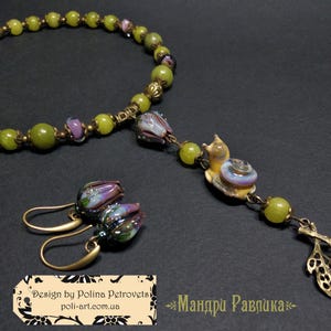 May include: A handmade jewelry set featuring a necklace and earrings. The necklace has green beads and a snail pendant. The earrings have purple and green flower-shaped beads. The set is made with gold-colored metal.