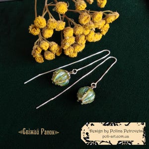 Puede incluir: Un par de pendientes de plata con cuentas de vidrio verde y blanco. Las cuentas tienen un diseño texturizado en forma de hoja. Los pendientes cuelgan de una cadena de plata.