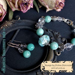 Può includere: Un set di gioielli con un braccialetto e orecchini. Il braccialetto e gli orecchini hanno perline turchesi e trasparenti, con dettagli in metallo color argento. Il braccialetto include anche una perla floreale decorativa. I gioielli sono esposti su una superficie scura.
