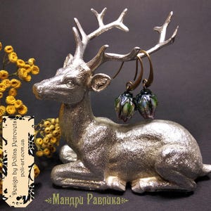 May include: A pair of handcrafted earrings with a green and purple floral design, suspended from bronze-colored hooks. The earrings are displayed on a silver-colored deer figurine. The background is dark, with yellow flowers and text.