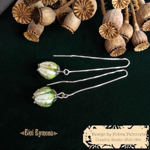 Op de afbeelding: Handgemaakte oorbellen met witte en groene bloemvormige kralen en zilveren kettingen. De oorbellen zijn geplaatst tegen een donkergroene achtergrond, met gedroogde zaaddozen op de achtergrond. Sierlijke en elegante oorbellen.